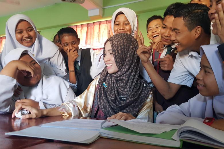 A young Asian female teacher wearing a head scarf smiles as she sits at a table in front of notebooks surrounded by smiling Asian boys and girls