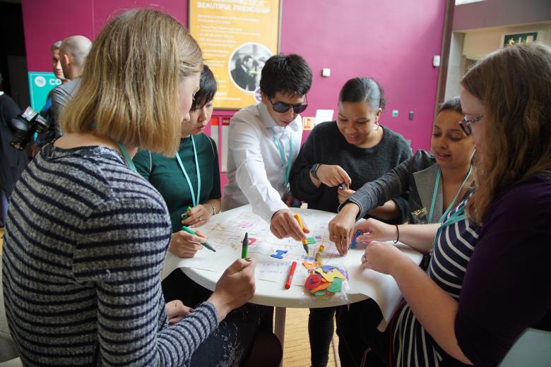 A diverse group of 6 adults lean over a table as they collaborate on a poster