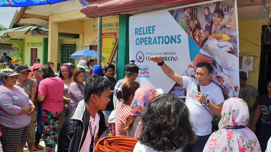 A group of people listen to man holding a microphone speaking in front of a large banner saying "RELIEF OPERATIONS"