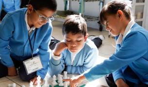 Two young boys and a young girl with brown hair and tan skin in blue school uniforms play chess on the floor