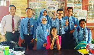 An Asian woman in a pink shirt smiles while seated at table surrounded by teenage students in blue uniforms