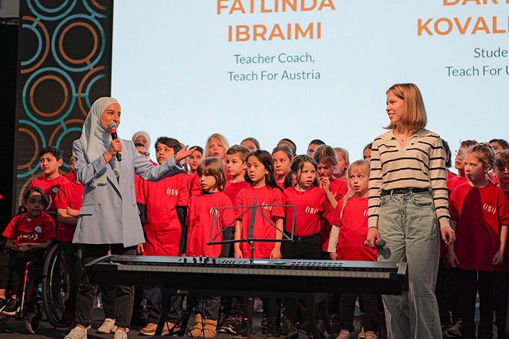 Two women, one wearing a head scarf and the other with blonde hair, stand in front of a choir of young children wearing red shirts