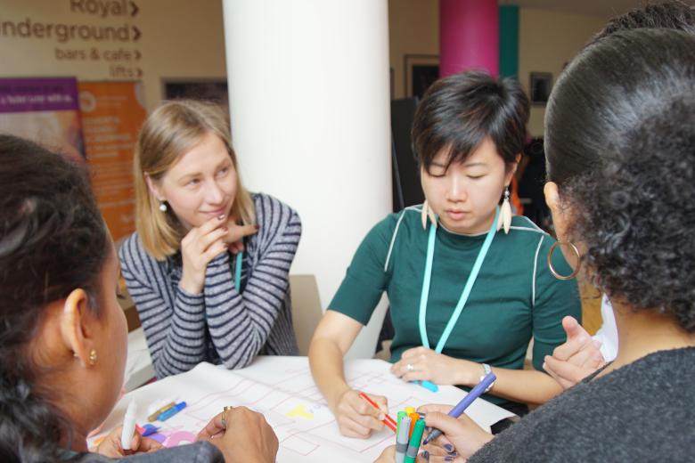 An Asian woman writes on chart on a table in front of her while a diverse group of women watch