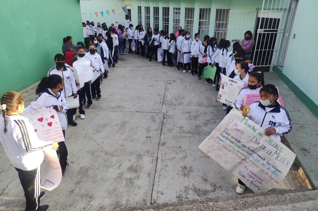 Teenage girls in white shirts holding big posters line two sides of an outdoor walkway between a light green wall and a white wall with windows