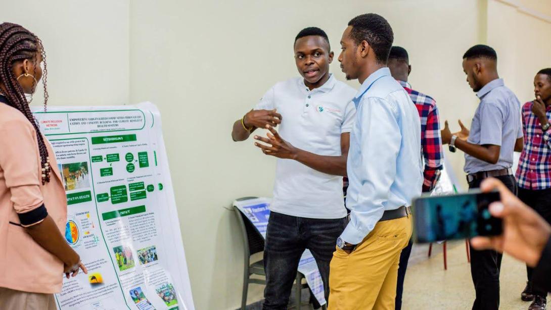 An African man stands in front of an academic poster explaining it to another young man while serveral other people surround them