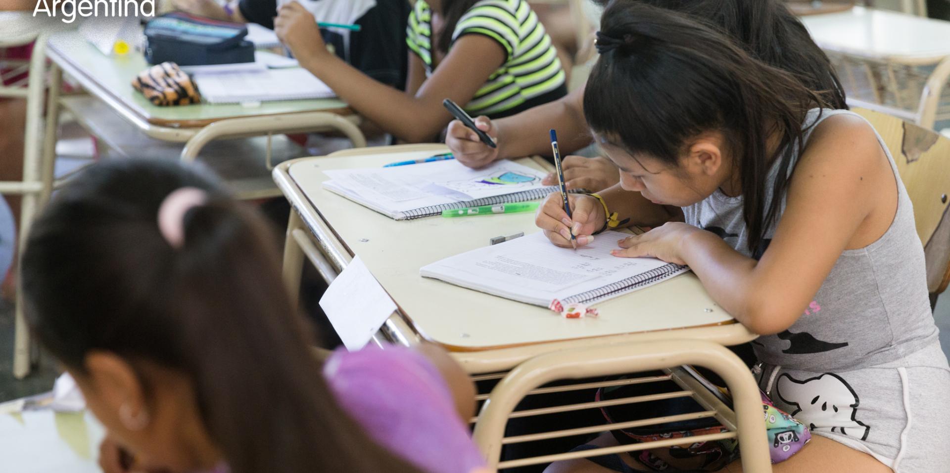 A young teen girl with a high ponytail writes in a notebook on her desk surrounded by classmates working at their desks