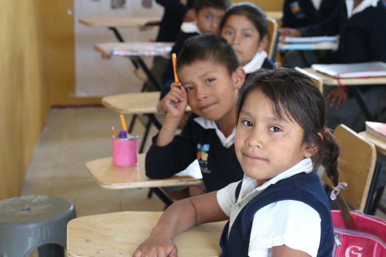 A brown-skinned little girl sits at a desk looking at the camera wtih a slight smile. Behind her is a boy at his desk also smiling and behind him is another girl.