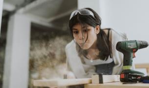A young teenage girl with brown hair and goggles on her head blows sawdust from a plant next to a powerdrill