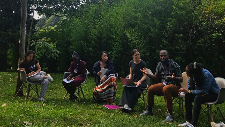 A group of six adults sit in a row on chairs outside on a green grass field in front of some trees