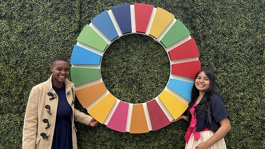 A young African girl and a young Peruvian girl stand on either side of a large SDG symbol mounted on a grass covered wall