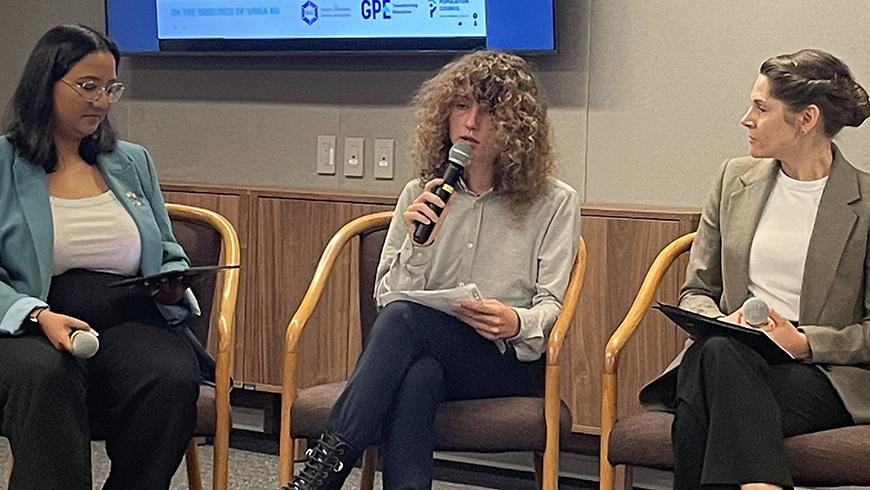 A young student with long curly blond hair wearing a button-down shirt and slacks sits in a chair speaking into a handheld microphone with two women in business casual outfits sit on either side of him listening to him