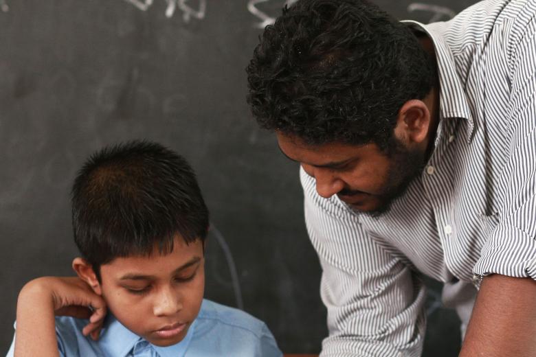 In front of a blackboard with writing on it, a male teacher with tan skin and brown hair leads over paper in front of a boy with similar coloring wearing a blue shirt