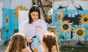A female teacher with long brown hair shows a globe to two female students we see from behind, with a backdrop of outdoor walls painted with sunflowers