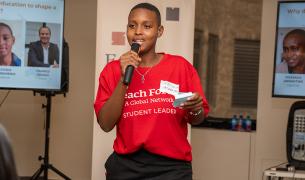A young african student speaks into a handheld microphone, she is wearing a red shirt that says "Teach For All student leader"