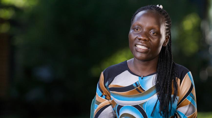 A woman with dark skin, long black braids and wearing a patterned top sits in front of a plain dark background