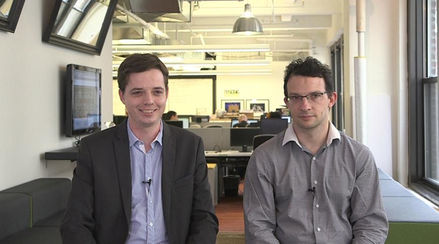 Two young white men with brown hair wearing button-downs sit next to each other facing the camera