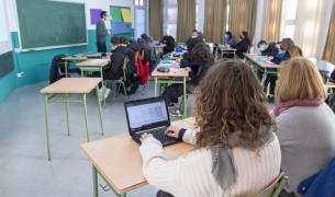 In the foreground, a woman uses a laptop computer while in the background a male teacher stands in front of a class of teenagers