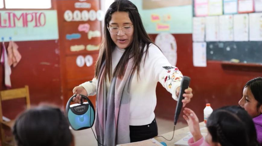 A young woman with long brown hair stands in a classroom speaking to young children