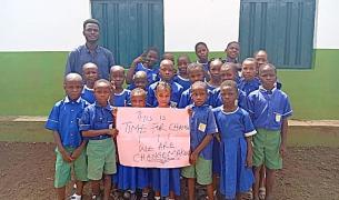 A group of African children in blue and green school uniforms and their male teacher stand together holding a sign that reads "This is Time for Change. We are Changemakers"