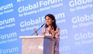 A young woman with long brown hair and tan skin stands at a podium in front of a background that says Global Forum for Shaping a Better Future