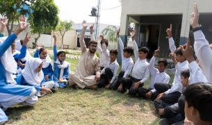 School children in uniform sit in a circle outside on grass with their teacher in the middle, all of them are raising one hands