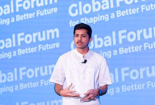 A young Indian man with a slight moustache and wearing a white shirt speaks in front of a blue background that says Global Forum for Shaping a Better Future