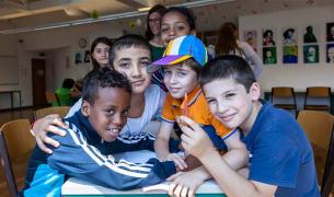 A group of boys of diverse ethnicities sitting at a table with notebooks crowd into the frame with their arms around each other smiling at the camera