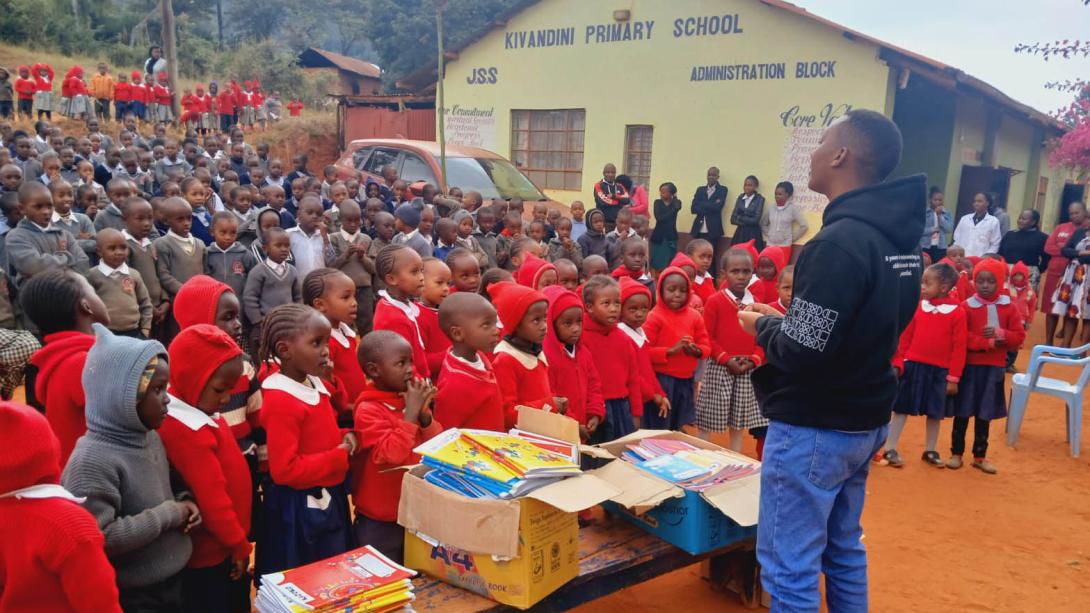 A young Black man stands in front of a bench piled with workbooks and a large crowd of Black children in school uniforms outside of a school