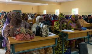 A room full of African women and men, many in colorful garments, sit at desks looking forward