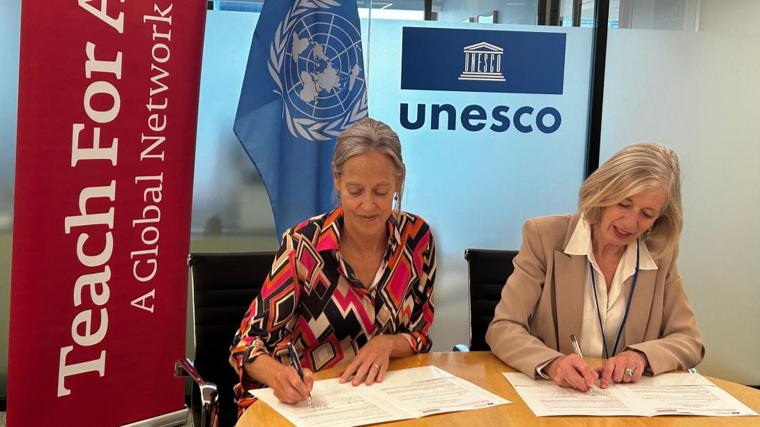 Two white women with greying blonde hair sit side by side signing papers in front of them, a red Teach For All banner on the right, and a UNESCO logo above one's head