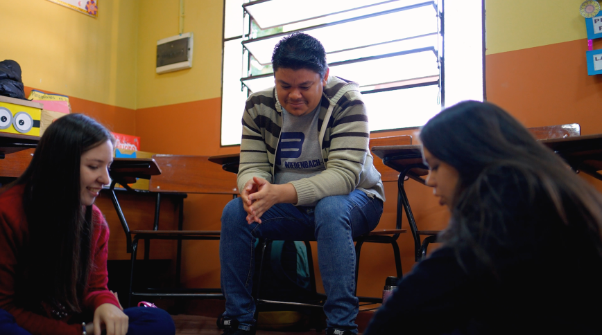 A young man with brown hair sits on a chair smiling while looking at something on the floor, where two young women are seated