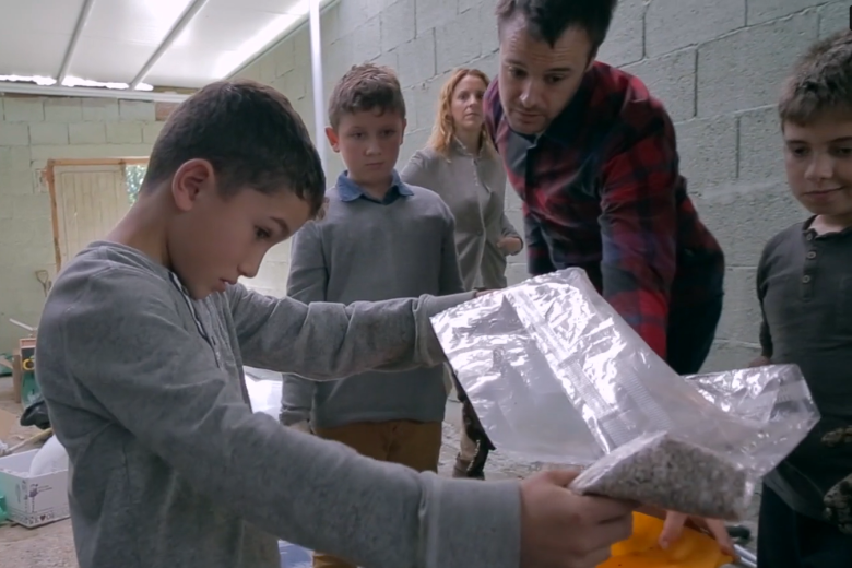A white boy holds a plastic bag as a young white man in a plaid shirt and three other white children lean in watching