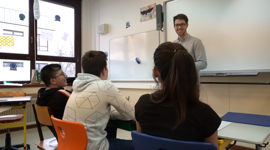 Through the backs of students' heads we see a young white male teacher with brown hair and glasses standing in front of a whiteboard.