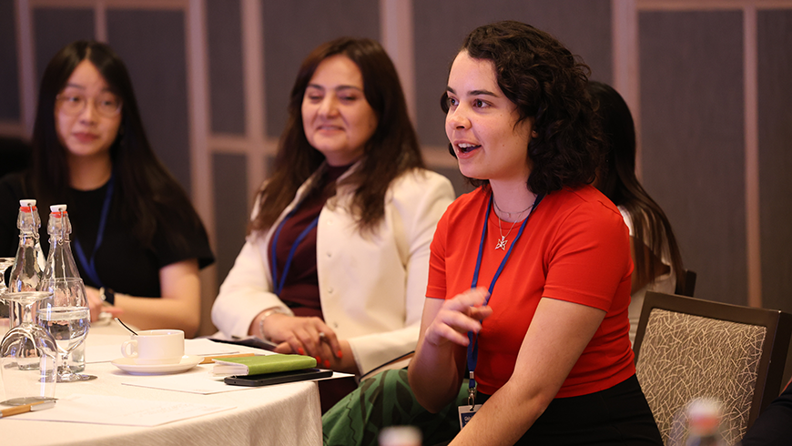 A young woman with shoulder-length brown curly hair and pale skin wearing a red shirt speaks while seated as others at the table watch and listen