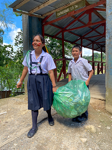 A girl and a boy with tan skin and brown hair wearing uniforms carry a bag of recycling