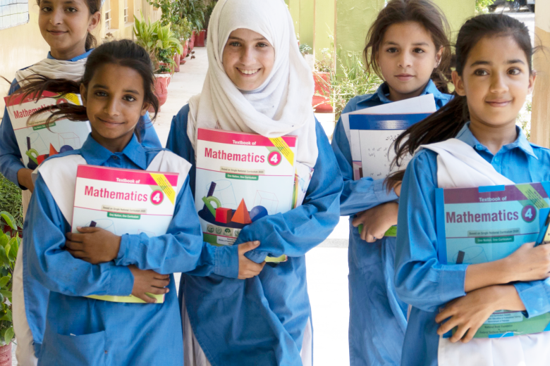 Five girls in blue dresses, one wearing a white head scarf, face the camera clutching mathematics workbooks